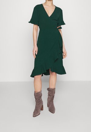 Vrouw draagt een donkergroene wikkeljurk met gerimpelde mouwen en zoom, gecombineerd met losse taupekleurige suède enkellaarzen met grove hakken.