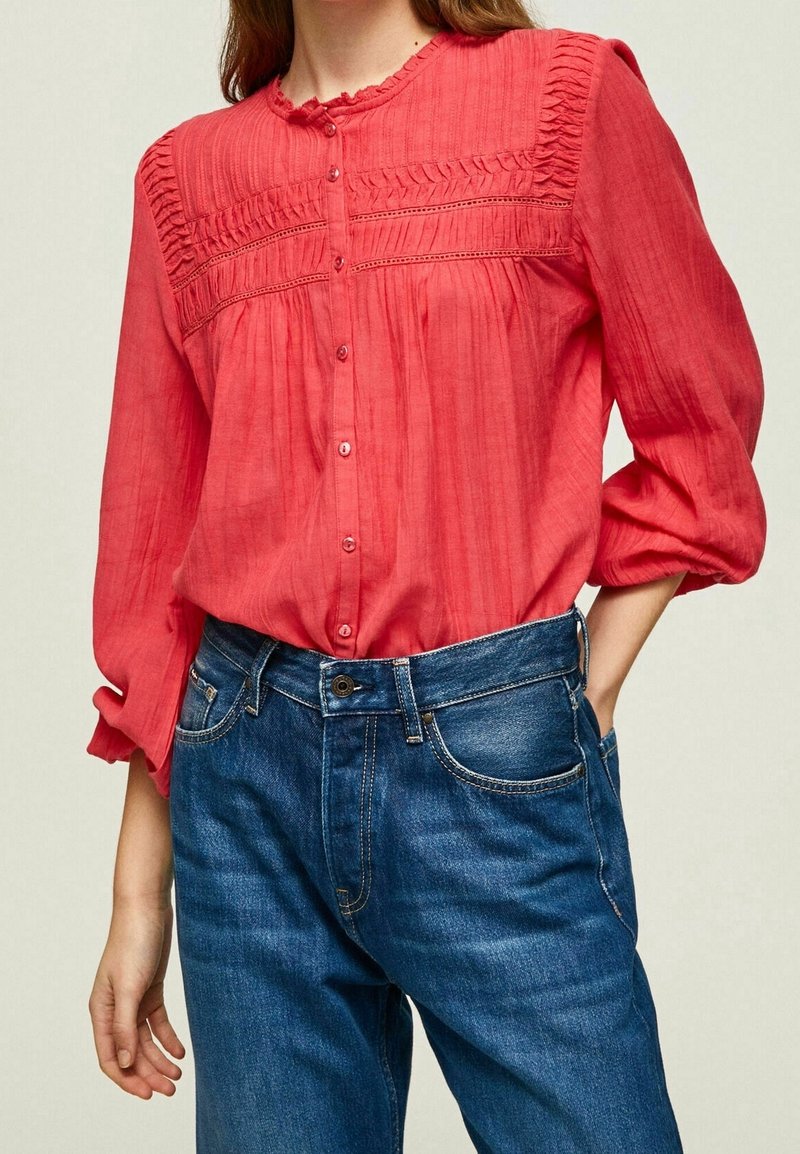 Femme portant un chemisier rouge à manches longues avec boutons sur le devant et plis détaillés, associé à un jean denim bleu taille haute.