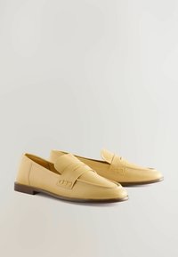 Gele leren loafers met een platte bruine zool, voorzien van een decoratieve band en stiksels op het bovenwerk voor extra textuur.