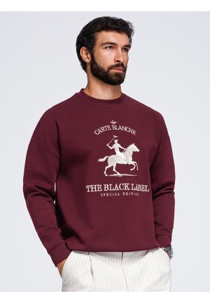 Mężczyzna z brodą ubrany w bordową bluzę z grafiką jeźdźca konnego i tekstem "Carte Blanche The Black Label Special Edition."
