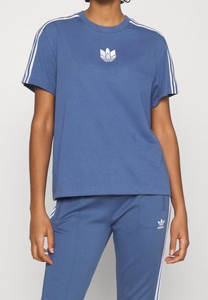 Person trägt blauen Adidas-Trainingsanzug mit weißen Streifen an Ärmeln und Hosen, mit Adidas-Logos auf Brust und Oberschenkel.