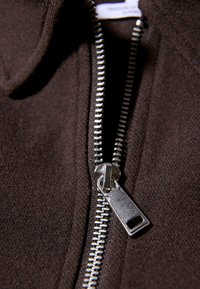 Close-up van een gedeeltelijk geopende zilveren rits op donkerbruin textuurstof, waarbij de details van de ritsen trekker en tanden zichtbaar zijn.