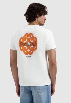 Man met krullend haar, die een wit T-shirt met een oranje abstract patroon en de tekst "Pure Path" draagt, en een blauwe spijkerbroek, zijdelings kijkend.