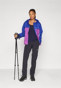 Lichtgewicht jas met kleurblokken in blauw en paars, gedragen over een donkere top. Zwarte outdoor broek, trekkingstokken en zwarte wandelschoenen.