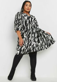 Svart- och vitmönstrad klänning med abstrakt mönster, puffärmar, v-ringning och utsvängd silhuett, bärs med svarta leggings och stövlar.