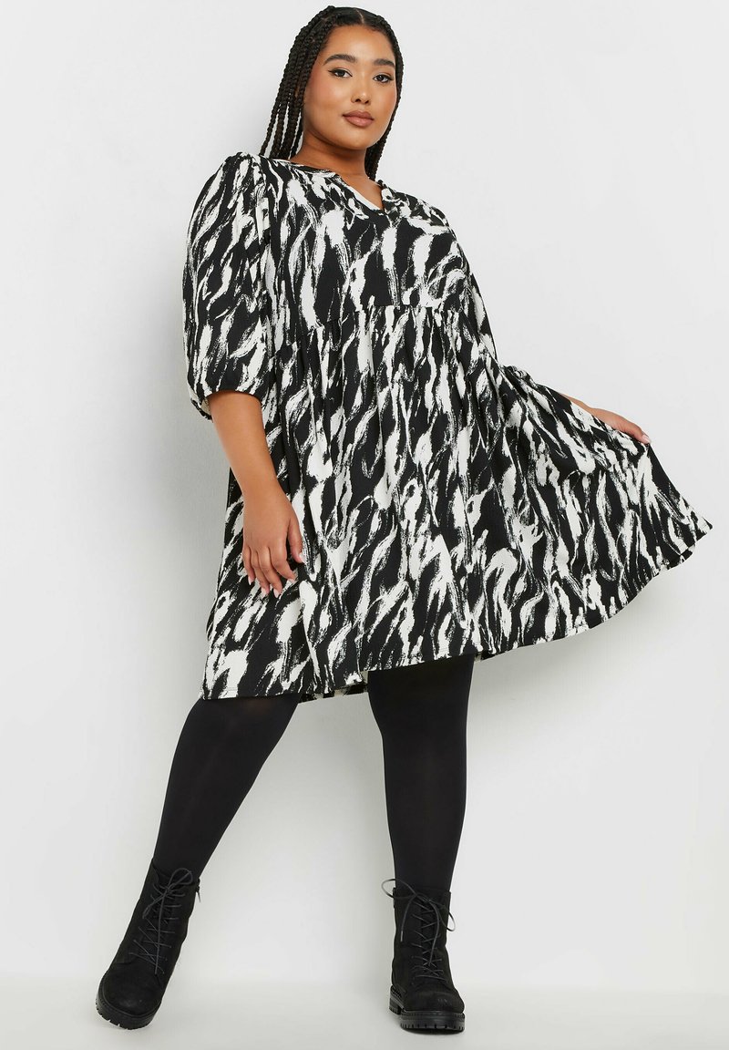 Svart- och vitmönstrad klänning med abstrakt mönster, puffärmar, v-ringning och utsvängd silhuett, bärs med svarta leggings och stövlar.