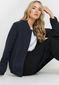 Giacca blu Navy con texture, colletto rotondo, maniche lunghe e polsini a costine, abbinata a pantaloni neri e un top bianco.