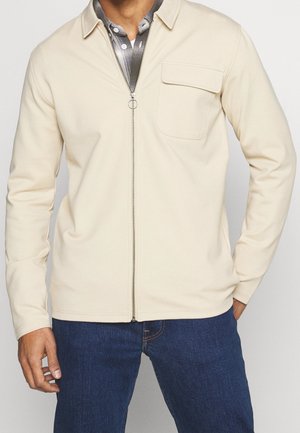 Chaqueta cremosa con cremallera y cuello, que presenta un bolsillo en el pecho y una textura suave, combinada con una camisa a cuadros debajo y jeans azules.