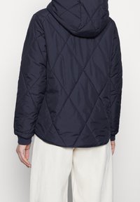 Granatowa pikowana kurtka z kapturem, z wzorem w romby, ściągaczami przy mankietach oraz gładką teksturą. Widok z tyłu nie pokazuje widocznych zapięć.