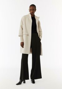 Beige oversized jas met een hoge kraag en knoopsluiting, gecombineerd met zwarte wijde broek en zwarte hoge laarzen. Gladde textuur en modern ontwerp.