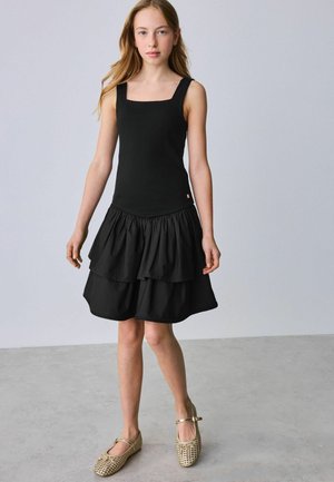 Junges Mädchen mit langen blonden Haaren, das ein ärmelloses schwarzes Kleid mit mehrlagigem Rock und goldenen Mary-Jane-Schuhen trägt und auf einem grauen Boden steht.