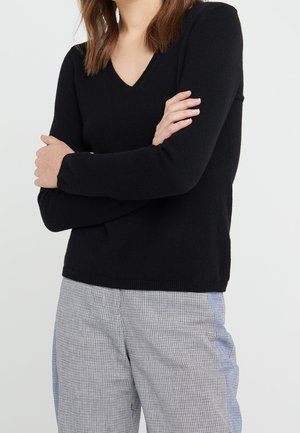 Person wearing a black V-neck long-sleeve sweater and gray checkered pants with crossed arms against a plain background.