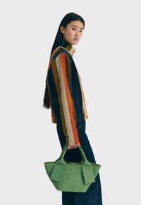 Una donna con lunghi capelli scuri indossa una giacca a righe e jeans scuri, mentre tiene una grande borsa verde su uno sfondo chiaro.