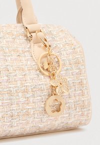 Μπεζ υφασμάτινη τσάντα με υφή και μεταλλικές κλωστές. Διακοσμημένη με χρυσό φυλαχτό που απεικονίζει μια πεταλούδα και κυκλικά στοιχεία προσαρμοσμένα πάνω της.