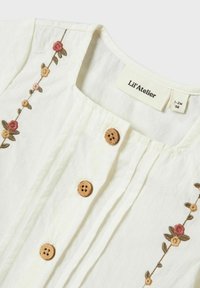 Top in cotone bianco con ricamo floreale in rosa e giallo. Ha un colletto rotondo, bottoni in legno e pieghe verticali. Etichetta del marchio visibile.