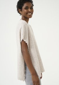 Beige gebreide poncho met korte mouwen, met een golvende zoom. Zachte textuur, comfortabele pasvorm en lichtgewicht ontwerp.