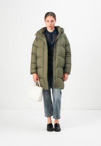 Vrouw in een lange olijfgroene pufferjas, doorzichtige marineblauwe top, cropped jeans, zwarte loafers, met een witte handtas tegen een witte achtergrond.