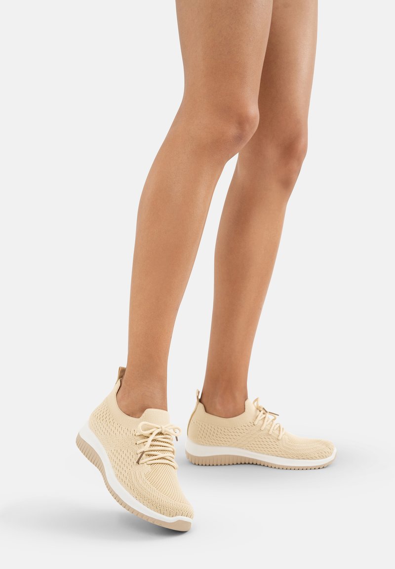 Beige gestrickte Sportschuhe mit niedrigem Design, strukturiertem Obermaterial, Schnürverschluss und weißer Gummisohle, mit einer Zuglasche an der Ferse.