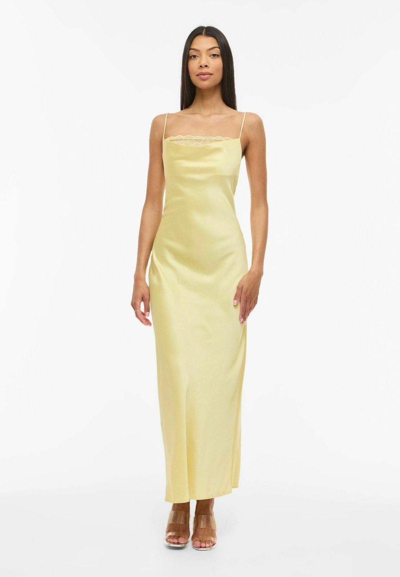 Femme aux longs cheveux foncés portant une robe nuisette en satin jaune avec des détails en dentelle et des talons à brides transparentes, se tenant devant un fond blanc.
