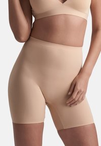 Donna che indossa pantaloni modellanti a vita alta di colore nude senza cuciture e un reggiseno abbinato, mano appoggiata delicatamente sulla coscia, sfondo neutro.