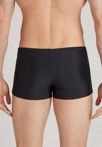 Schwarze Badehorts aus glattem Material, mit einem enganliegenden Design, elastischem Bund und minimalen Nähten, von hinten betrachtet.