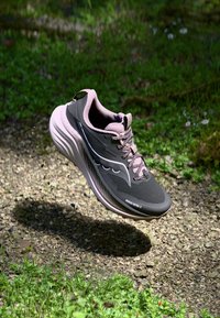 Zapatilla deportiva en negro y gris con acentos en rosa claro, parte superior de malla, gruesa mediasuela blanca y suela texturizada, flotando sobre una superficie de grava.