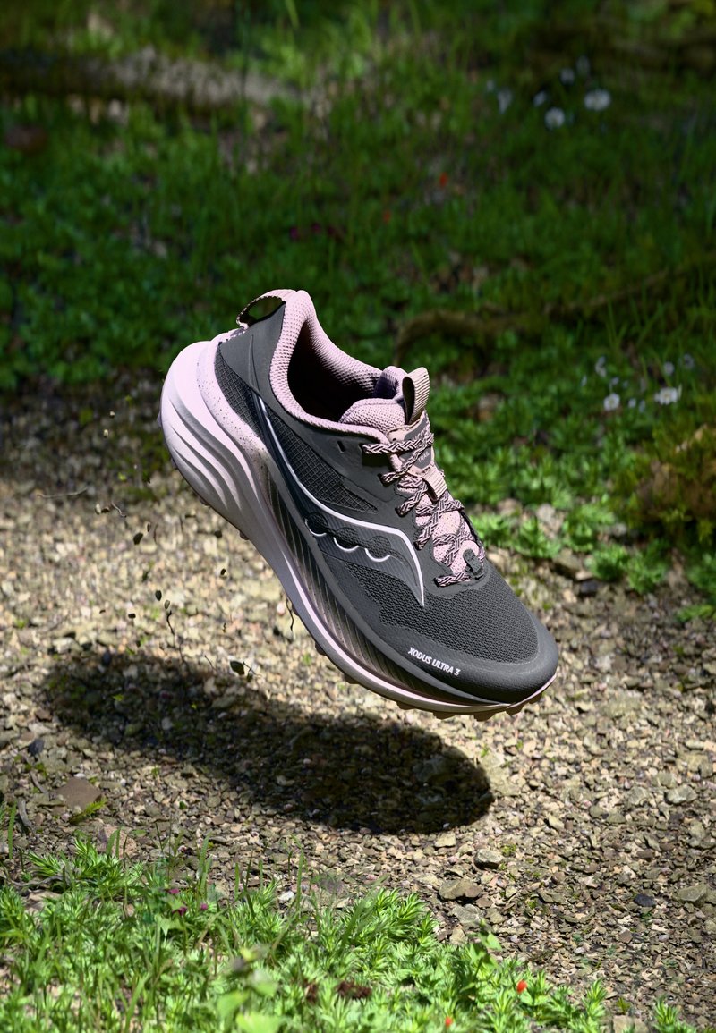 Zapatilla deportiva en negro y gris con acentos en rosa claro, parte superior de malla, gruesa mediasuela blanca y suela texturizada, flotando sobre una superficie de grava.