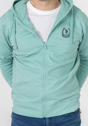 Hombre con sudadera con cremallera color verde azulado claro con el logo de U.S. Polo Assn. sobre camiseta blanca y vaqueros azules, con las manos atrás.