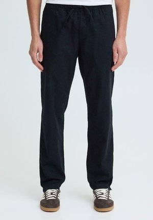 Man wearing loose black drawstring pants and brown sneakers with white laces, standing against a plain white background.