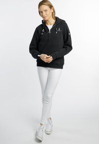 Zwarte hoodie met voorkant rits, trekkoord en wit ankerlogo. Gecombineerd met witte skinny jeans en witte sneakers. Glad stofgevoel.