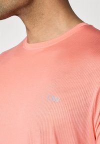Tricou ușor, texturat, de culoare coral, cu guler rotund și detalii subtile cu logo în colțul din dreapta jos.