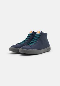Paire de baskets montantes bleu marine avec des lacets bleu sarcelle, semelles grises et logo orange sur des étiquettes blanches sur la languette, légèrement inclinées vers la droite.