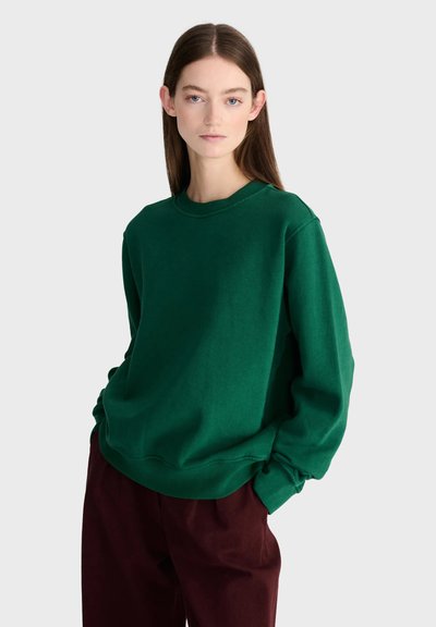 Jeune femme aux longs cheveux bruns portant un sweat-shirt vert et un pantalon rouge foncé, debout les mains dans les poches devant un fond uni.