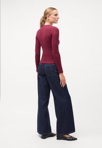 Top burgundiu cu mâneci lungi asortat cu blugi largi de culoare închisă și pantofi negri fără toc. Topul are un design ajustat și o textură netedă.