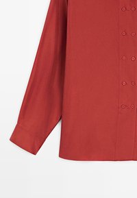 Red button-up shirt with long sleeves, smooth texture, and a straight cut. Features six matching buttons along the front and a cuffed sleeve.
