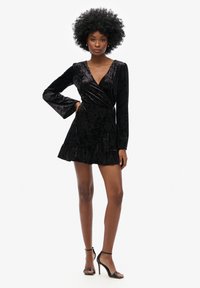 Robe portefeuille en velours noir avec des manches longues, un décolleté en V plongeant, des poignets évasés et un ourlet volé, assortie à des sandales à talons hauts noires.