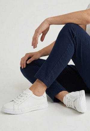 Osoba siedząca na podłodze, ubrana w granatowe spodnie i białe trampki na sznurowadła, z jedną nogą zgiętą i jedną ręką opartą na kolanie.