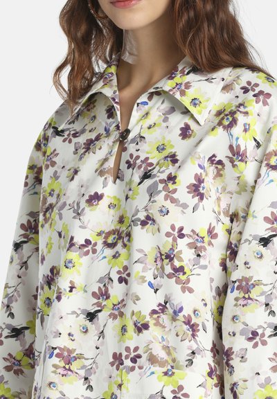 Femme portant un chemisier blanc à manches longues avec un grand col, motif floral en jaune, violet et beige, boutonné au niveau du col.