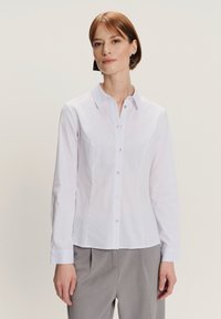Donna che indossa una camicia bianca aderente con maniche lunghe e pantaloni grigi, in piedi contro uno sfondo chiaro e semplice.