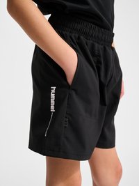 Personne portant un short noir à taille élastique avec les mains dans les poches, arborant le logo blanc « hummel performance » sur la jambe gauche.