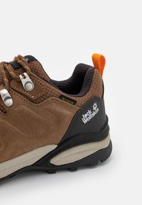 Brauner Wanderschuh aus Wildleder und Mesh-Materialien, graue Gummisohle, orangefarbener Ziehschlaufe, reflektierende Akzente und das Markenlogo auf der Seite sichtbar.
