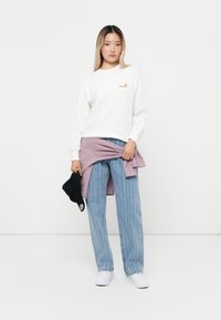 Hvid langærmet skjorte med lille orange logo, sammen med lyseblå stribede jeans. En lilla skjorte bundet om taljen og en sort kasket.