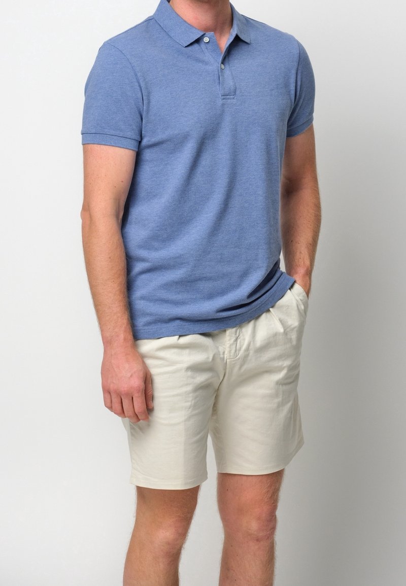 Homme portant un polo bleu clair à manches courtes et un short beige jusqu'aux genoux, une main dans la poche, sur fond blanc.