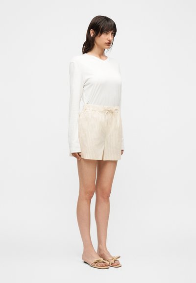 Γυναίκα στέκεται φορώντας λευκό μακρυμάνικο τοπ, μπεζ σορτς με ζωνάκι και διακριτικές ρίγες, καθώς και μπεζ σανδάλια με κόμπο σε λευκό φόντο.