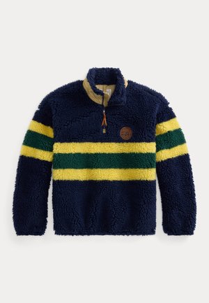 Personne portant une polaire bleu marine avec des rayures jaunes et vertes, une chemise à carreaux, un pantalon camouflage, des chaussures vertes, une écharpe à carreaux, un bonnet en tricot beige et des lunettes de soleil foncées.