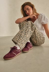 Burgunderfarbene Wildledersneaker mit weißen Akzenten und Schnürsenkeln, kombiniert mit hellen Hosen im Leopardenmuster, auf einer flachen Oberfläche getragen.