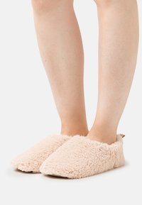 Light pink fluffy slippers with a soft, textured exterior and rounded toe design, featuring an open back and minimalistic brown tab detail.