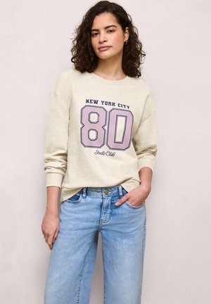 Donna con capelli ricci che indossa una felpa beige con la scritta "New York City 80 Studio Club" e jeans azzurri, in posa su uno sfondo neutro.