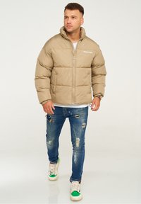 Beige Daunenjacke mit hohem Kragen, Reißverschluss und gestepptem Design. Kombiniert mit verwaschenen blauen Jeans und mehrfarbigen Sneakers.