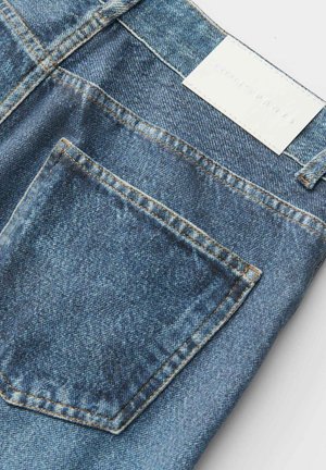 Nahaufnahme der Gesäßtasche einer blauen Jeans mit sichtbaren Nähten und einem weißen, rechteckigen Markenschild am Bund.